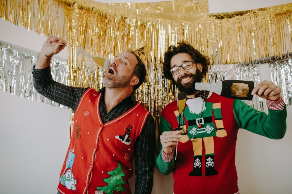 Two men enjoy a playful holiday party with festive sweaters and props, surrounded by tinsel decorations.